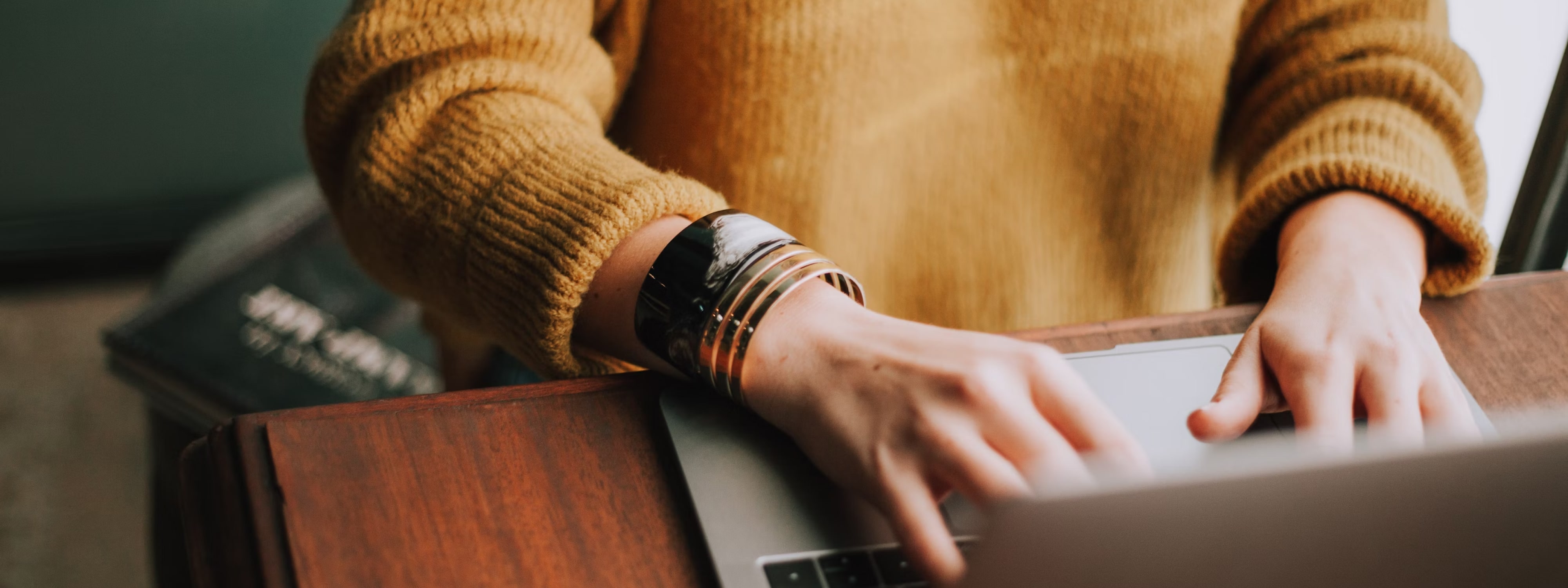 Person in mustard sweater typing on laptop at wooden desk