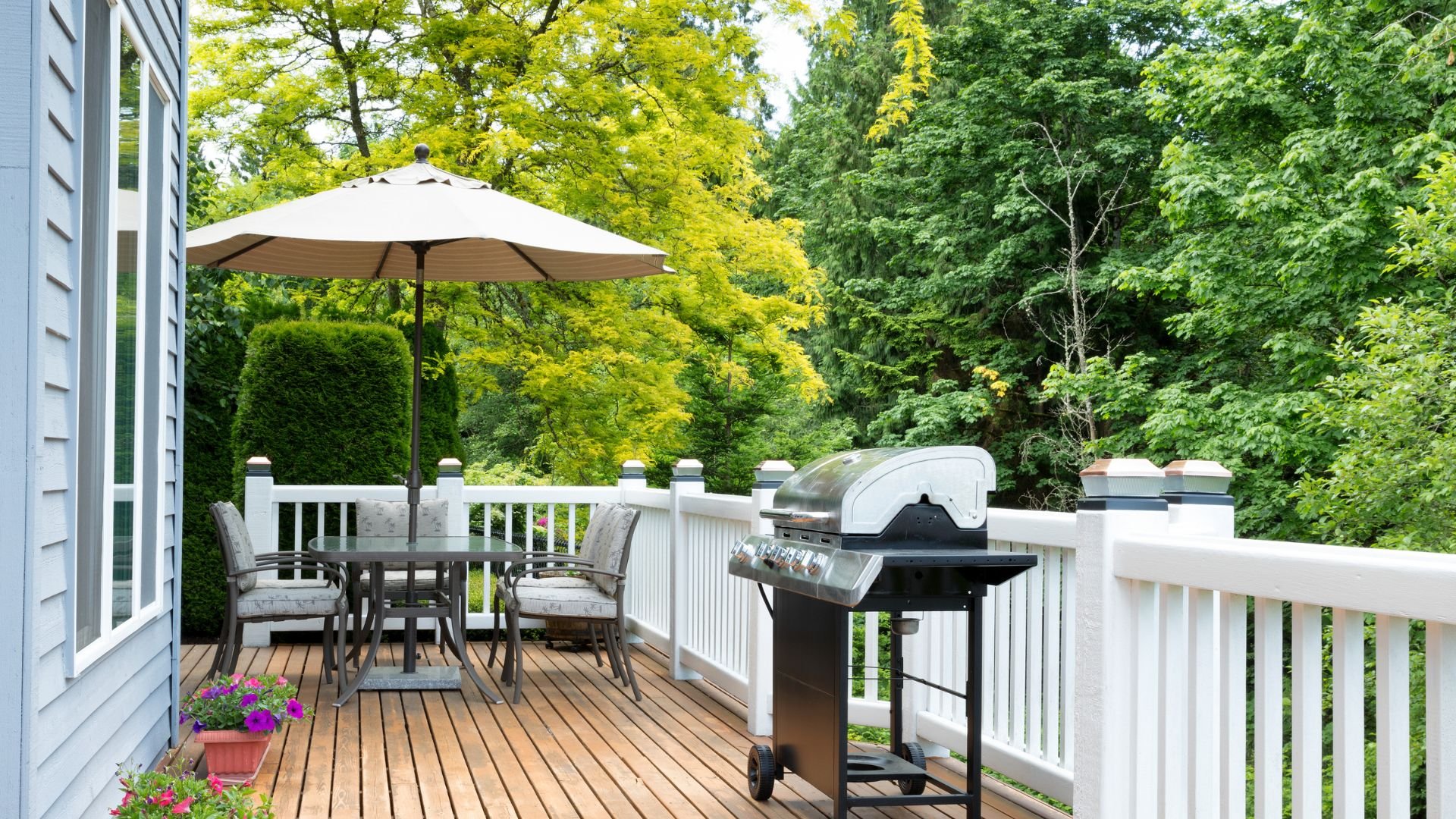 Wooden deck with patio furniture, umbrella, grill, and lush green trees